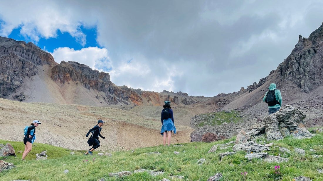 People hiking on a grassy mountain with rocky cliffs and a cloudy sky.