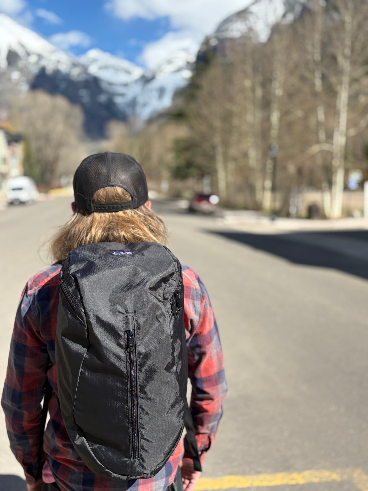 Person wearing a black Jagged Edge daypack on a road with mountains in the background